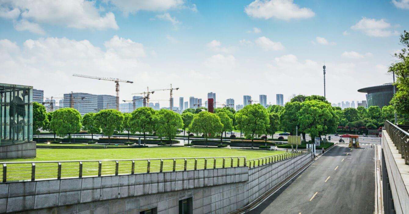 A view of a vibrant green park with trees to indicate Malaysia Urban Renewal Projects, surrounded by construction sites and modern skyscrapers under a bright blue sky with clouds.